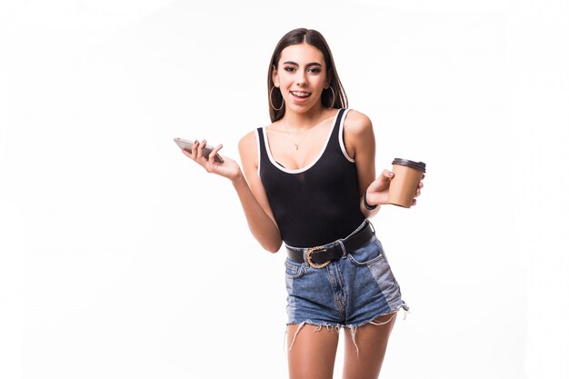 Joven modelo tierna se queda con teléfono y taza de café frente a la pared blanca