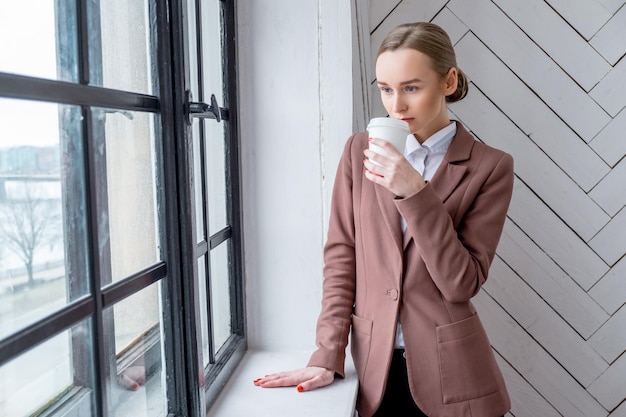 Joven moderna con una chaqueta marrón bebiendo café cerca de la ventana de una habitación.