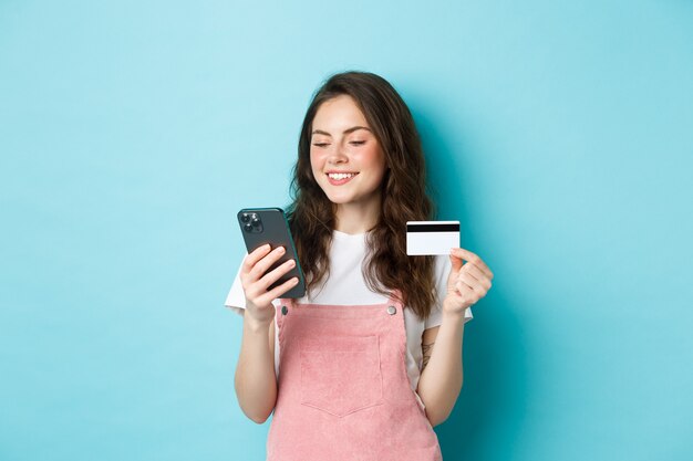 Joven moderna pagando en línea con tarjeta de crédito, escriba información en el teléfono inteligente, mirando la pantalla con cara de satisfacción, comprando en la aplicación, de pie sobre fondo azul.