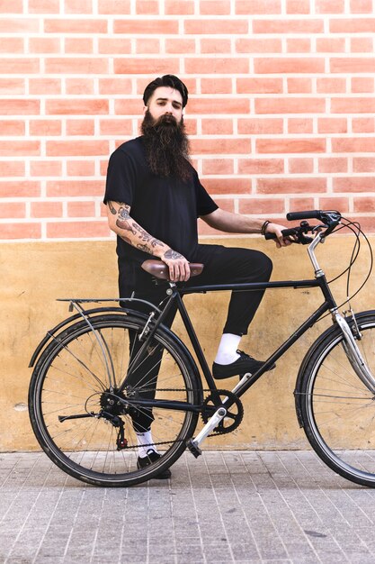 Joven moderno de pie con su bicicleta contra la pared de ladrillo