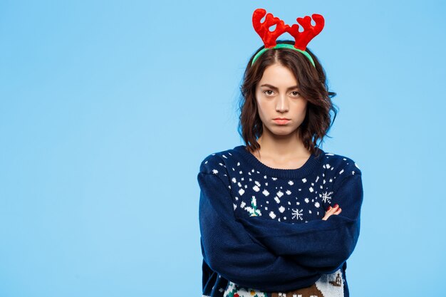 Joven molesta hermosa chica morena en suéter de punto y astas de renos de Navidad sobre pared azul
