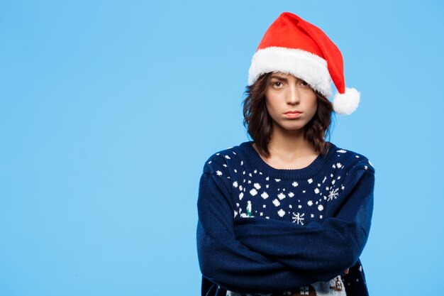 Joven molesta hermosa chica morena en suéter de punto y sombrero de navidad sobre pared azul