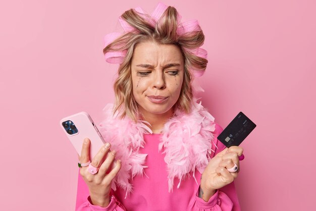 Una joven molesta y seria mira la tarjeta bancaria que sostiene la aplicación de descargas de teléfonos inteligentes para el pago en línea hace que el peinado se prepare para la fiesta viste un vestido festivo aislado sobre fondo rosa