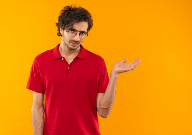 Joven molesto en camisa roja con gafas ópticas apunta al lado con la mano aislada en la pared naranja