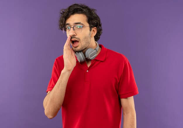 Joven molesto en camisa roja con gafas ópticas y con auriculares tiene la mano en la boca pretendiendo llamar a alguien aislado en la pared violeta