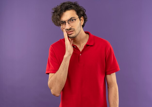 Joven molesto en camisa roja con gafas ópticas pone la mano en la cara aislada en la pared violeta