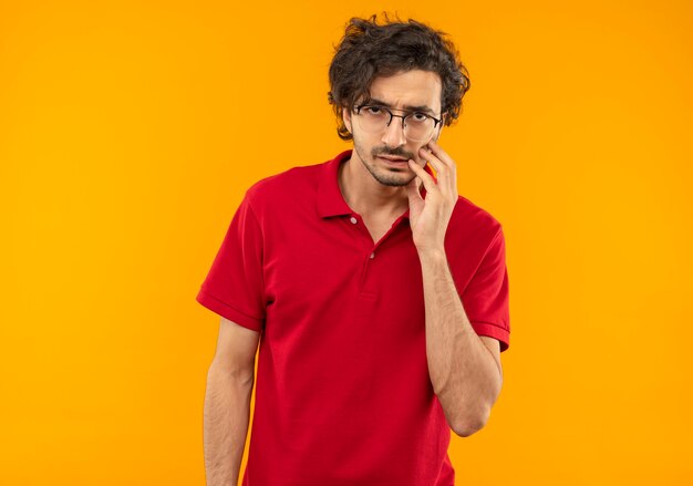 Foto gratuita joven molesto en camisa roja con gafas ópticas pone la mano en la cara y parece aislado en la pared naranja