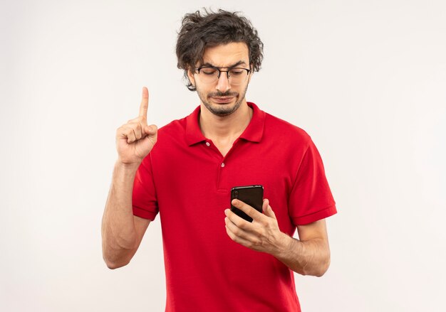 Joven molesto en camisa roja con gafas ópticas sostiene el teléfono y apunta hacia arriba aislado en la pared blanca