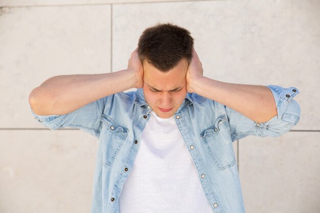 Joven molesto cubriendo las orejas con las manos en la pared al aire libre