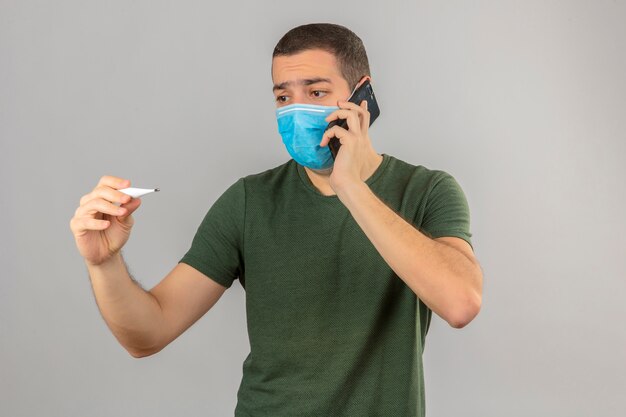 Joven molesto hombre en máscara médica mirando el termómetro digital y hablando por smartphon aislado en blanco