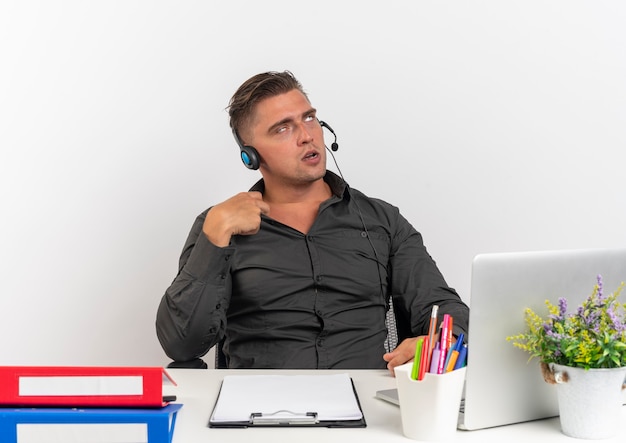 Joven molesto rubio oficinista hombre en auriculares se sienta en el escritorio con herramientas de oficina usando laptop rodando ojos tiene collar aislado sobre fondo blanco con espacio de copia