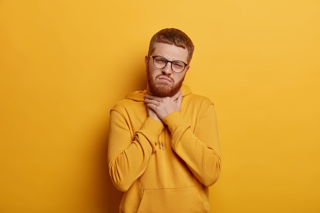 El joven molesto tiene el pelo corto y pelirrojo, se toca el cuello y sufre de dolor de garganta, siente dolor al tragar, usa una sudadera con capucha, aislado sobre una pared amarilla. Mal síntoma