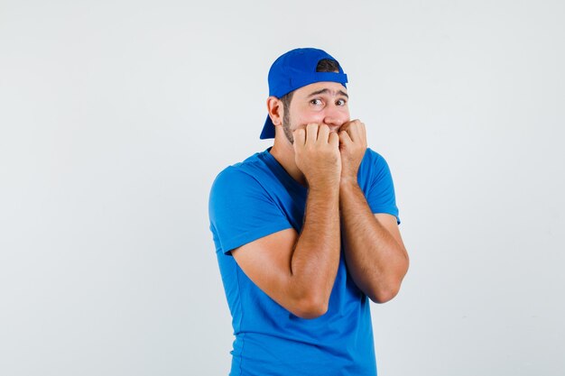 Joven mordiendo los puños emocionalmente en camiseta azul y gorra y mirando asustado