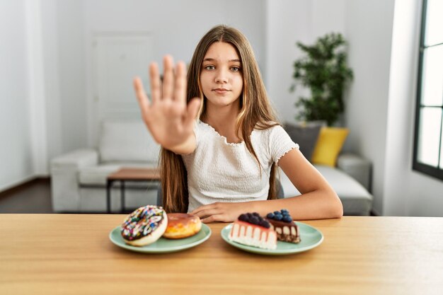 Joven morena adolescente comiendo pasteles y dulces con la mano abierta haciendo la señal de alto con gesto de defensa de expresión seria y segura