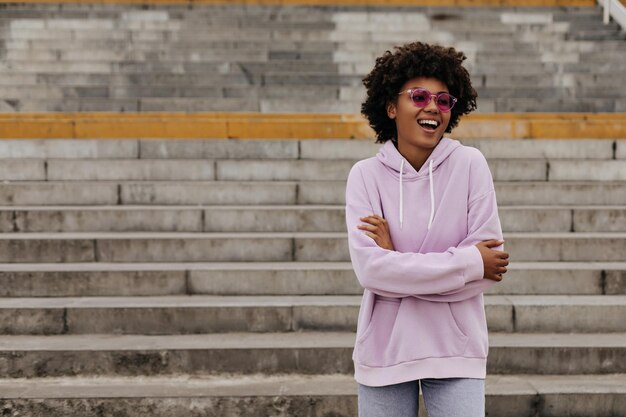 Una joven morena de buen humor, de piel oscura y rizada, con gafas de sol rosas y una elegante sudadera con capucha morada, sonríe, cruza los brazos y posa cerca de las escaleras