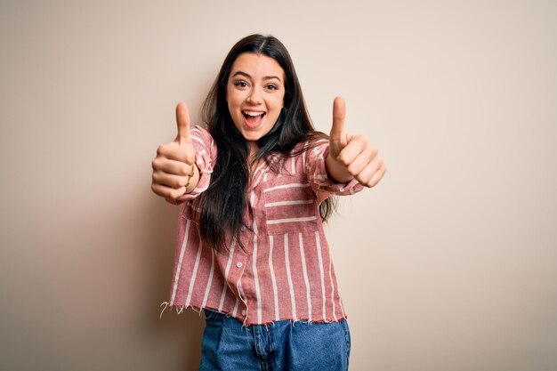 Joven morena con camisa casual a rayas sobre un fondo aislado aprobando hacer un gesto positivo con los pulgares hacia arriba sonriendo y feliz por el éxito Gesto ganador