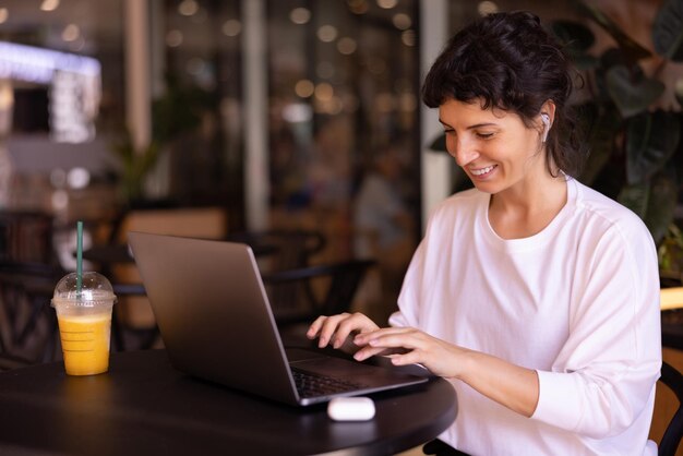 Una joven morena caucásica positiva con camisa trabaja como redactora escribiendo en una laptop sentada en un café durante el día
