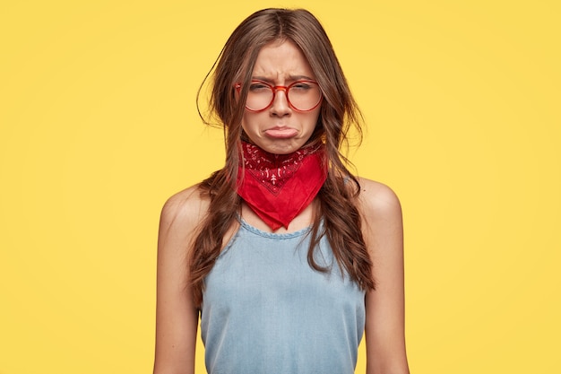 Joven morena deprimida con gafas posando contra la pared amarilla