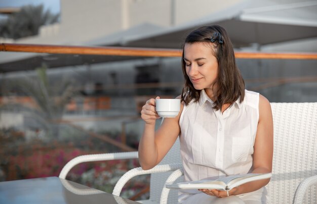 Joven morena disfruta de la mañana con una taza de bebida caliente y un libro en sus manos. Concepto de descanso y relajación.