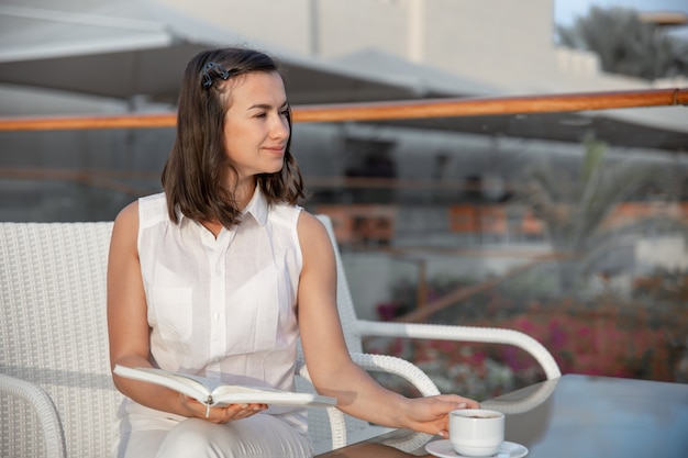 Joven morena disfruta de la mañana con una taza de bebida caliente y un libro en sus manos.