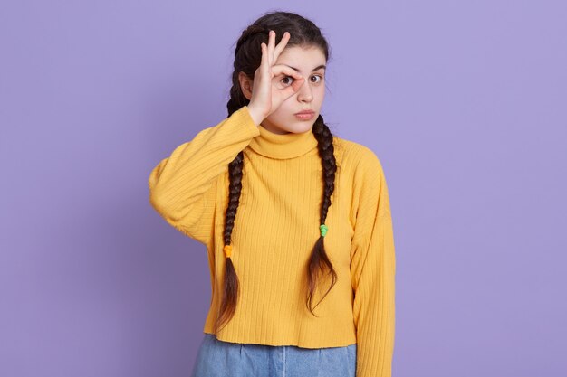 Joven morena empujando el signo ok y cubriendo su ojo con él, posando aislado sobre la pared de color lila