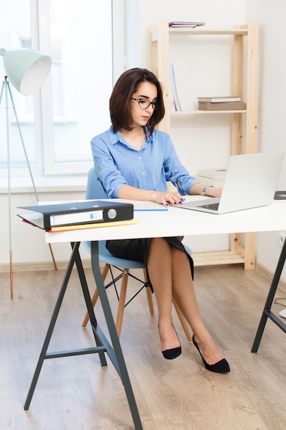 Una joven morena está sentada a la mesa en la oficina. Viste camisa azul y zapatos negros. Ella está escribiendo en la computadora portátil.