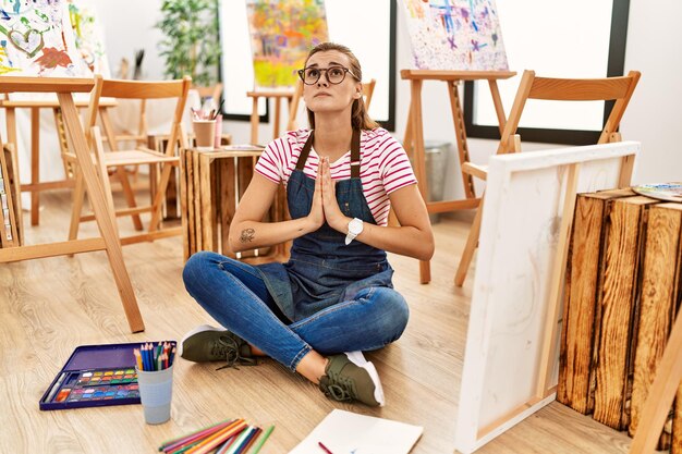 Joven morena en el estudio de arte sentada en el suelo rogando y rezando con las manos juntas con expresión de esperanza en la cara muy emocional y preocupada rogando