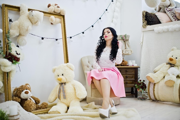 Joven morena con falda rosa y blusa blanca posada en el interior contra la habitación con oso de juguete