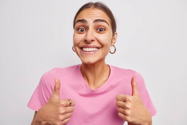 Una joven morena feliz sonríe alegremente, mantiene los pulgares hacia arriba y muestra un signo de aprobación satisfecho con el servicio. La buena retroalimentación dice que es excelente, vestida con una camiseta rosa casual aislada sobre el fondo blanco.