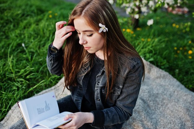 Una joven morena con jeans sentada en una manta contra un árbol de flores de primavera y lee el libro