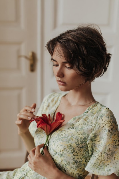 Joven morena de pelo corto con un elegante vestido verde y amarillo se ve triste y sostiene una gran flor roja La dama posa cerca de una puerta blanca de madera