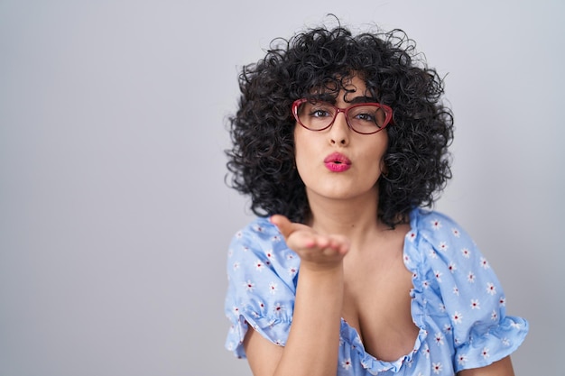 Foto gratuita joven morena con el pelo rizado y gafas sobre un fondo aislado mirando a la cámara soplando un beso con la mano en el aire siendo encantadora y sexy. expresión de amor.