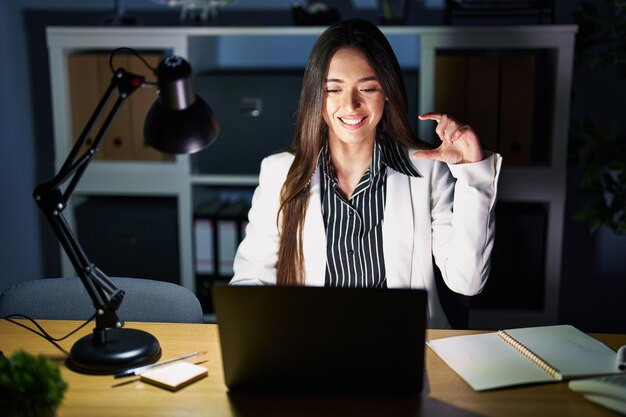 Joven morena que trabaja en la oficina por la noche con una laptop sonriendo y confiada gesticulando con la mano haciendo un signo de tamaño pequeño con los dedos mirando y el concepto de medida de la cámara