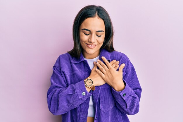 Joven morena con ropa informal sonriendo con las manos en el pecho con los ojos cerrados y un gesto agradecido en la cara. concepto de salud