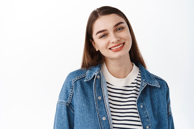 Joven morena sonriente con dientes perfectos mirando al frente, de pie relajado contra la pared blanca en ropa casual