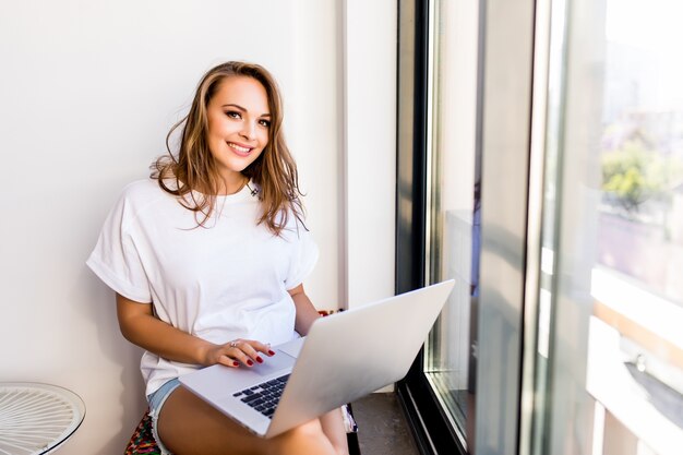 Joven morena sonriente está sentada en una silla moderna cerca de la ventana en una habitación luminosa y acogedora en casa. Ella está trabajando en una computadora portátil en un ambiente relajante.