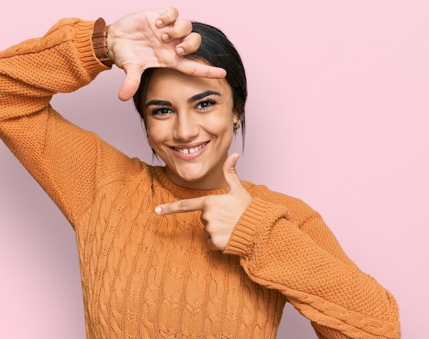 Joven morena con suéter casual de invierno sonriendo haciendo marco con manos y dedos con cara feliz. concepto de creatividad y fotografía.