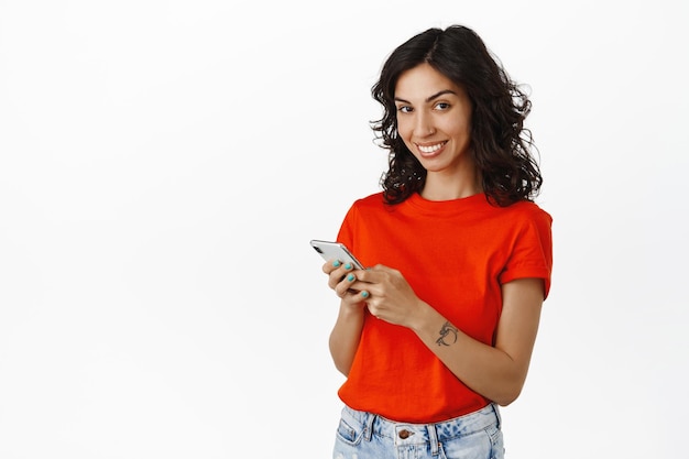 Joven morena usando teléfono móvil, sosteniendo en las manos un teléfono inteligente y mirando feliz a la cámara, sonriendo mientras chatea, usando la aplicación, de pie sobre fondo blanco