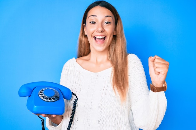 Foto gratuita joven morena usando teléfono vintage gritando orgullosa celebrando la victoria y el éxito muy emocionada con los brazos levantados