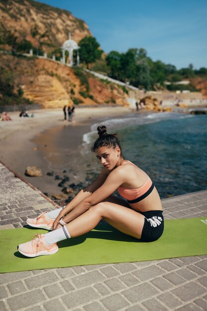 Joven morena vistiendo traje de fitness, estirando sobre estera de yoga al aire libre en el muelle en verano. Chica en forma, haciendo posturas de yoga por el mar.