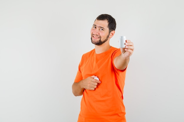 Joven mostrando una botella de píldoras en camiseta naranja y mirando alegre, vista frontal.