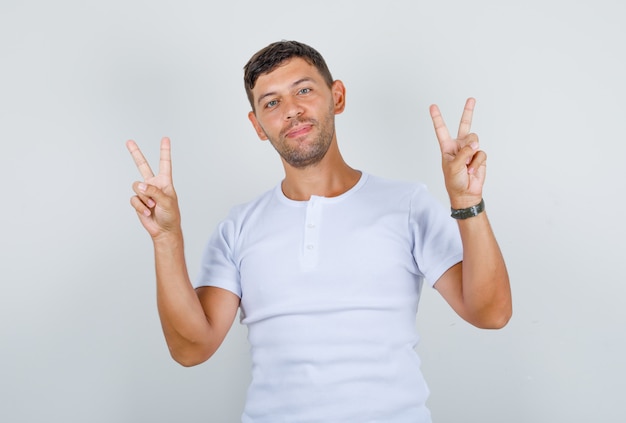 Joven mostrando los dedos haciendo el signo de la victoria en camiseta blanca y mirando feliz, vista frontal