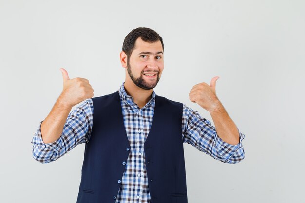 Foto gratuita joven mostrando doble pulgar hacia arriba en camisa, chaleco y mirando feliz