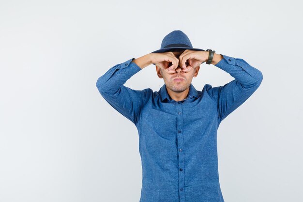 Joven mostrando gesto de gafas en camisa azul, sombrero y mirando enfocado. vista frontal.