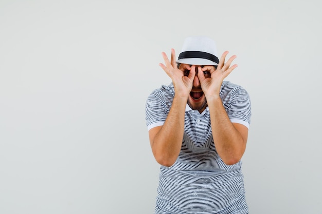 Foto gratuita joven mostrando gesto de gafas en camiseta, sombrero y mirando divertido, vista frontal.