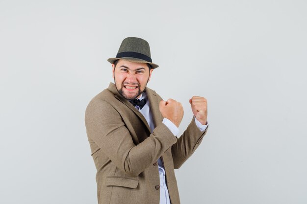 Joven mostrando gesto de ganador en traje, sombrero y con suerte, vista frontal.