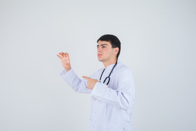 Joven mostrando gesto de garra, apuntando hacia el lado izquierdo con uniforme de médico y mirando alegre