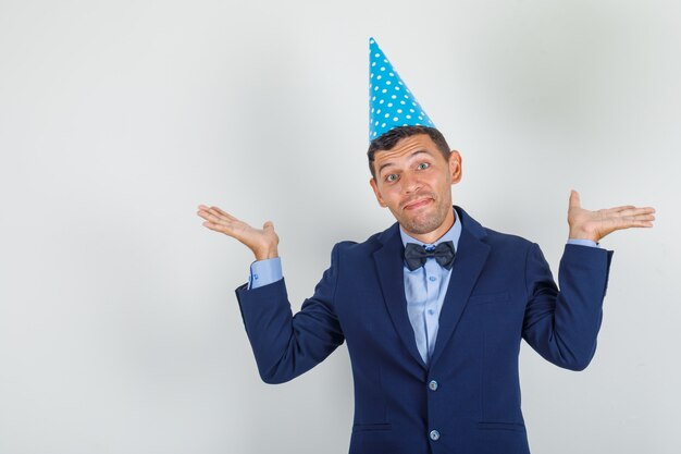 Joven mostrando gesto de impotencia en traje, sombrero de fiesta azul y mirando confundido