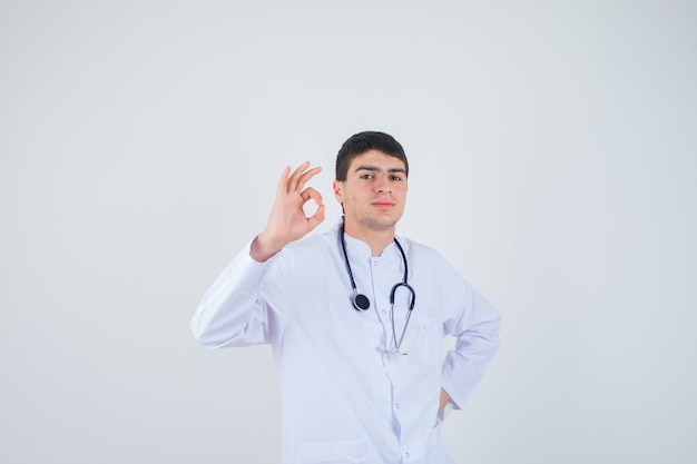 Joven mostrando gesto ok en uniforme de médicoy mirando confiado