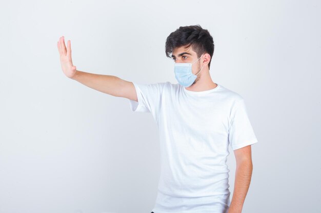 Joven mostrando gesto de parada en camiseta blanca, máscara y mirando confiado